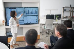 Financial professional presenting agency staffing data on an office screen — tracking APS and ACT Government voluntary redundancy rounds.