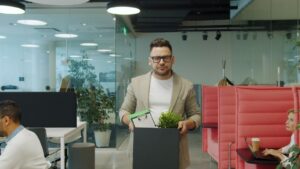 A man in a jacket carrying a box of personal belongings as he leaves an open-plan office after being made redundant