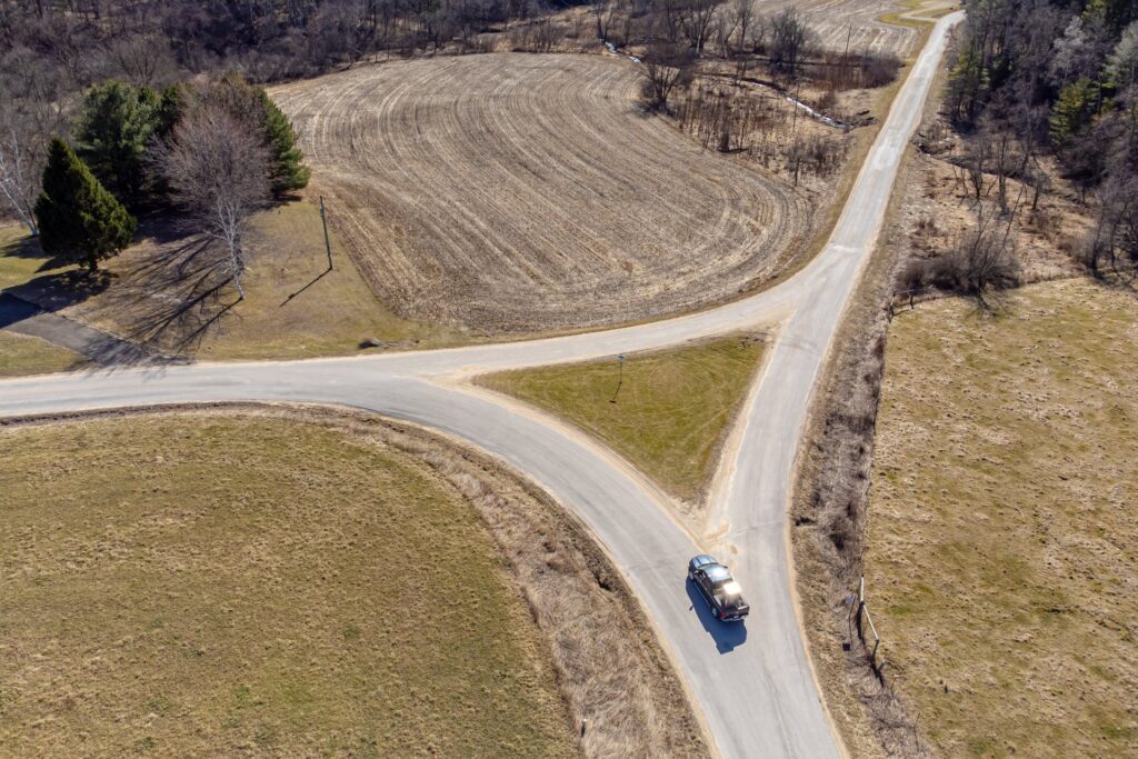 Aerial view of a car on a country road approaching a fork, with one road curving left and the other running straight ahead between fields.